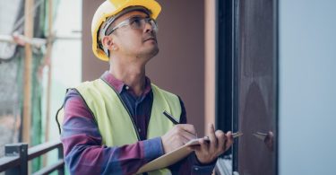 A man in a hard hat is holding a clipboard and is using a pencil to mark things down on the clipboard.