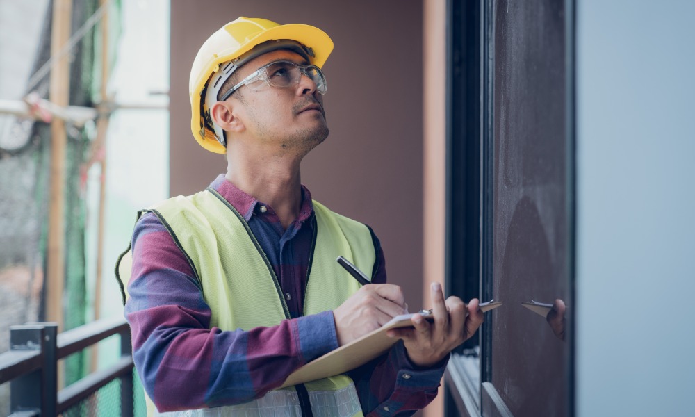 A man in a hard hat is holding a clipboard and is using a pencil to mark things down on the clipboard.