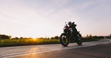 A rider wearing a black helmet on a red motorcycle driving on a concrete road next to a green field during a sunset.