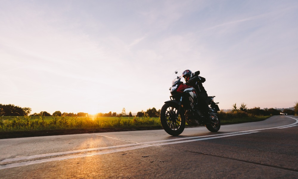 A rider wearing a black helmet on a red motorcycle driving on a concrete road next to a green field during a sunset.