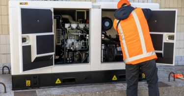 A rear view of a man in a blue hoodie and orange high-visibility vest working on a large generator with the panels open.