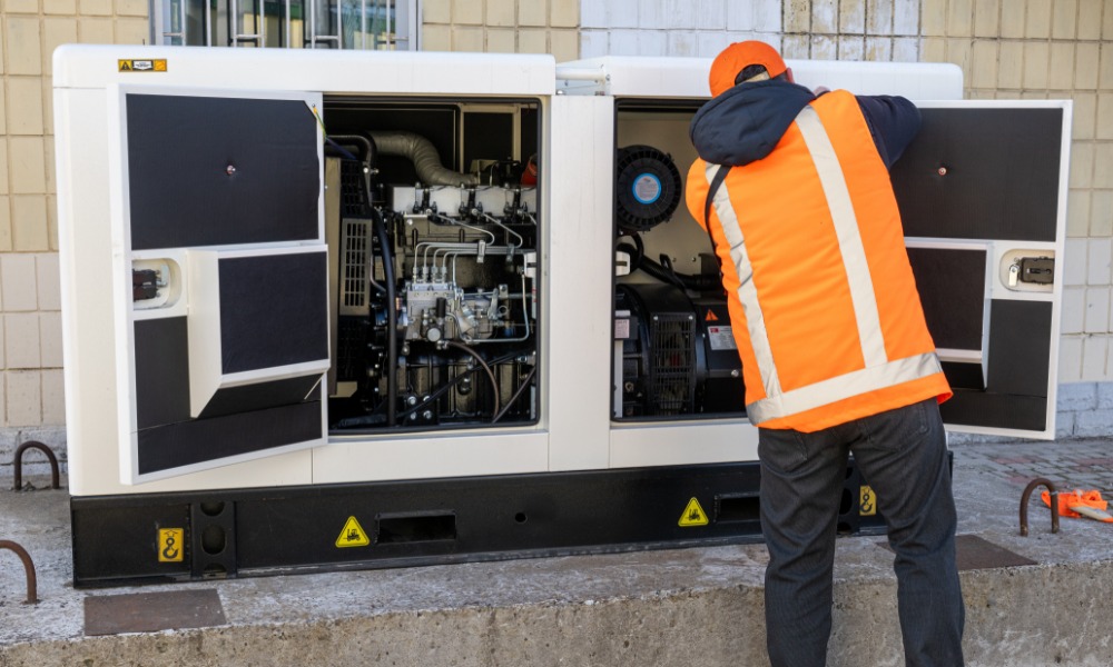 A rear view of a man in a blue hoodie and orange high-visibility vest working on a large generator with the panels open.