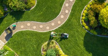 Person pushes a lawn mower across a manicured yard with sweeping paths, stepping stones, and colorful garden beds.