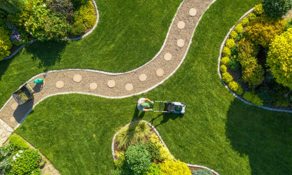 Person pushes a lawn mower across a manicured yard with sweeping paths, stepping stones, and colorful garden beds.