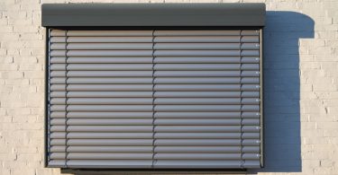 A black security shutter covers a single window of a building. The building has a tan brick wall.