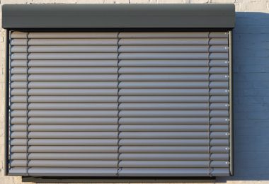A black security shutter covers a single window of a building. The building has a tan brick wall.