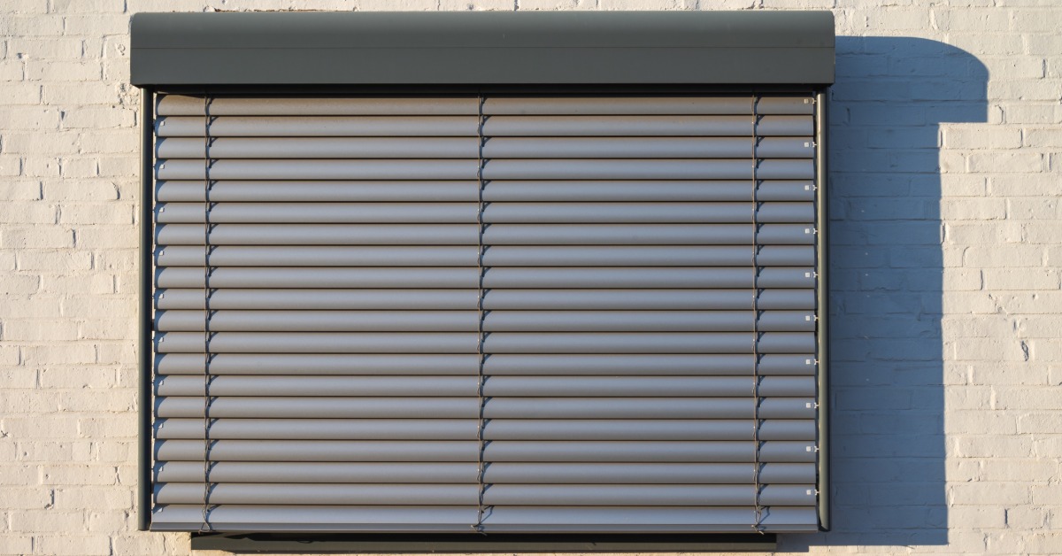 A black security shutter covers a single window of a building. The building has a tan brick wall.