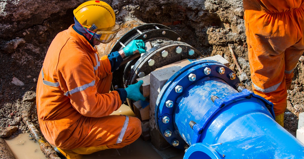 Two workers wearing orange uniforms and yellow helmets looking at a large pipe with bolts in a hole underground.