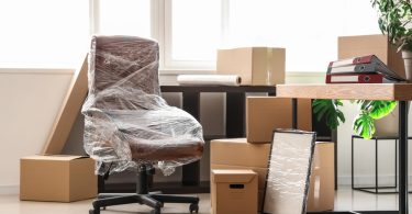 A brown office chair wrapped in clear plastic, cardboard moving boxes, a wrapped frame, and a desk sit in an office.