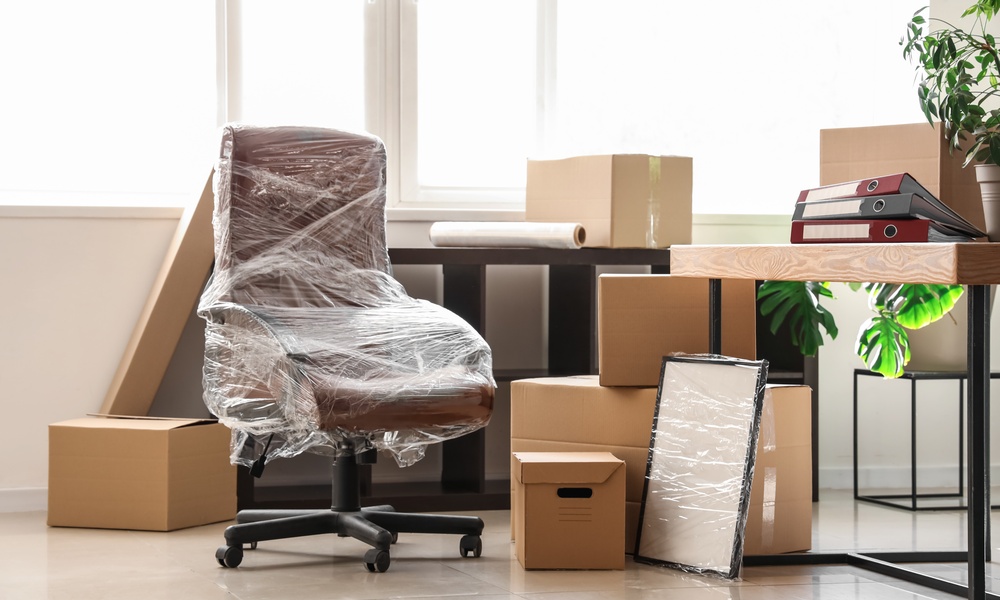 A brown office chair wrapped in clear plastic, cardboard moving boxes, a wrapped frame, and a desk sit in an office.