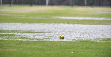 A yellow softball with red stitching sits in a large rain puddle on a grassy area. Rain is still falling.