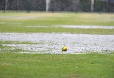 A yellow softball with red stitching sits in a large rain puddle on a grassy area. Rain is still falling.