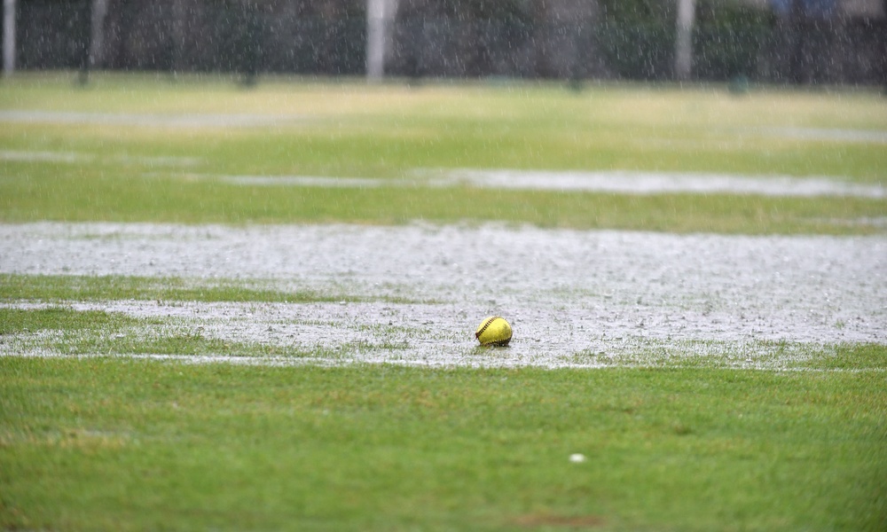 A yellow softball with red stitching sits in a large rain puddle on a grassy area. Rain is still falling.
