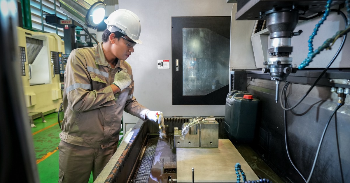 A man wearing a white hard hat and a tan jumpsuit uses a spotlight to look at the inside of a CNC machine.