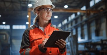 An employee wearing a white hard hat and safety glasses in an industrial facility. The employee holds a tablet.