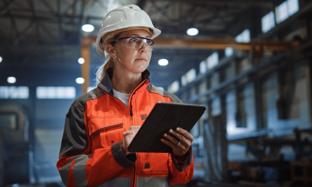 An employee wearing a white hard hat and safety glasses in an industrial facility. The employee holds a tablet.