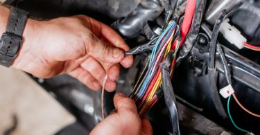 A mechanic wearing a black wristwatch moving cables attached to a car’s wiring harness in a bright room.