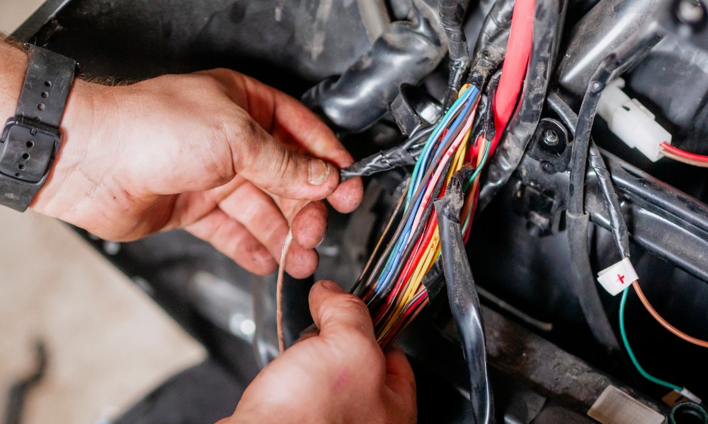 A mechanic wearing a black wristwatch moving cables attached to a car’s wiring harness in a bright room.