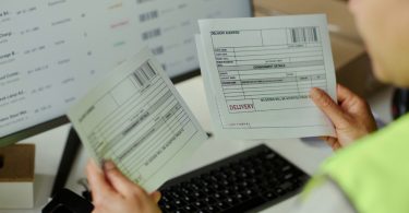 An employee compares printed inventory records with information displayed on a laptop screen in a warehouse office.