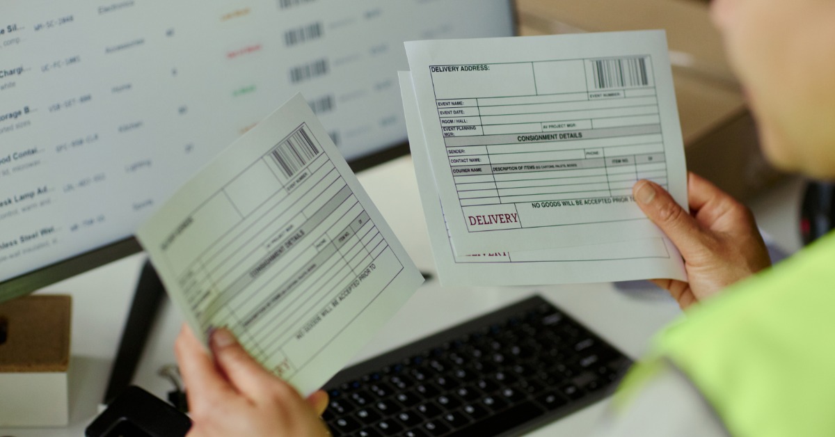 An employee compares printed inventory records with information displayed on a laptop screen in a warehouse office.