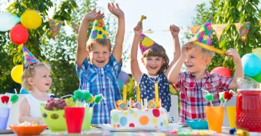 Four kids wearing colorful party hats and smiling around a small cake on a table at an outdoor birthday party.