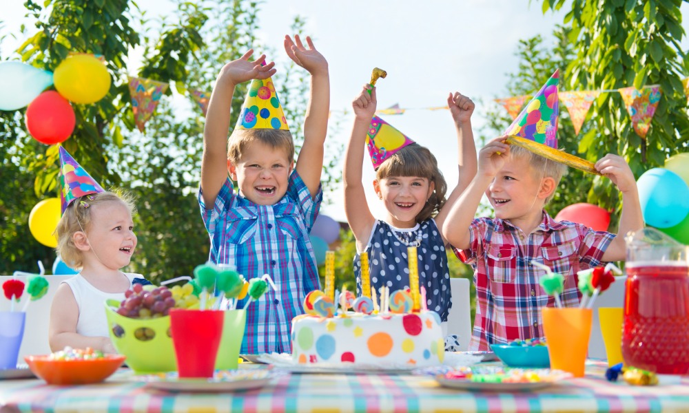 Four kids wearing colorful party hats and smiling around a small cake on a table at an outdoor birthday party.