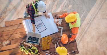 Three construction workers wearing hard hats outside lean over a pile of wood planks to read blueprints.