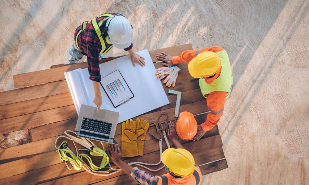 Three construction workers wearing hard hats outside lean over a pile of wood planks to read blueprints.