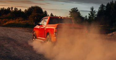 A bright red pickup truck driving down a dirt road next to a forest. The sun sets in the background.