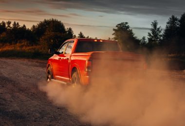 A bright red pickup truck driving down a dirt road next to a forest. The sun sets in the background.