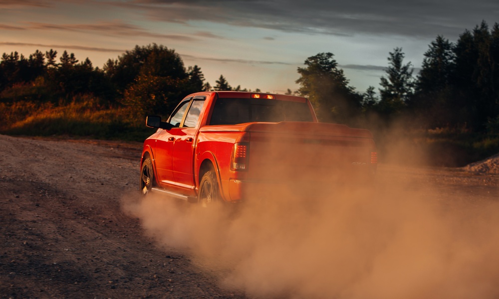 A bright red pickup truck driving down a dirt road next to a forest. The sun sets in the background.