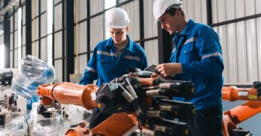 Two employees wearing hard hats inspect orange robotic arms in an industrial facility. One technician holds a tablet.