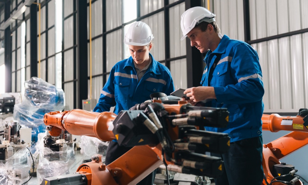 Two employees wearing hard hats inspect orange robotic arms in an industrial facility. One technician holds a tablet.