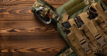 A green protective vest with attached pouches resting on a brown wooden surface beside folded green fabric.