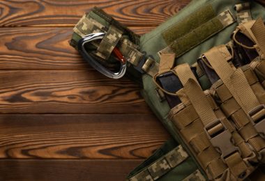 A green protective vest with attached pouches resting on a brown wooden surface beside folded green fabric.