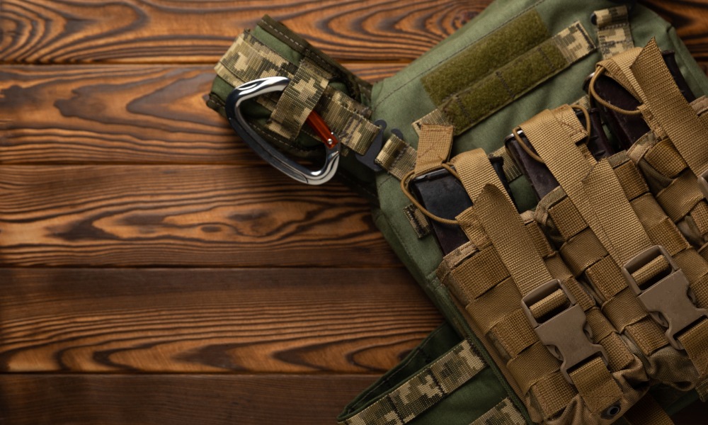 A green protective vest with attached pouches resting on a brown wooden surface beside folded green fabric.