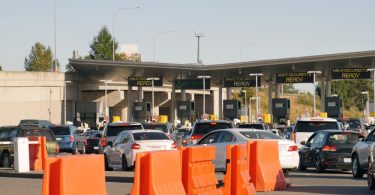 Cars line up at a busy border checkpoint with document-ready signs above booths and orange barriers in front.