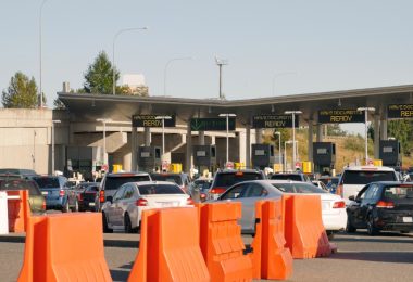 Cars line up at a busy border checkpoint with document-ready signs above booths and orange barriers in front.