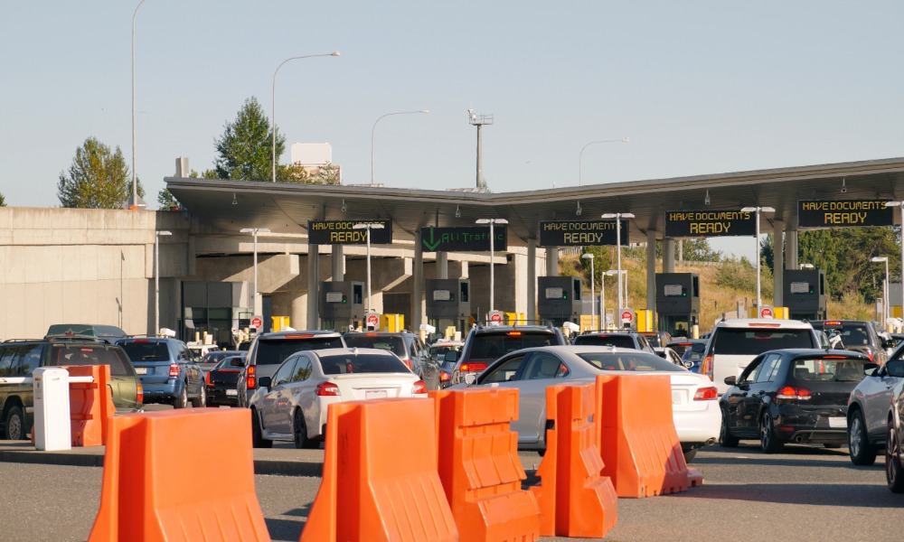 Cars line up at a busy border checkpoint with document-ready signs above booths and orange barriers in front.