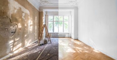 An empty room split between worn plaster walls with a ladder and renovated white walls with a herringbone wood floor.