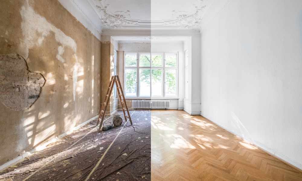 An empty room split between worn plaster walls with a ladder and renovated white walls with a herringbone wood floor.