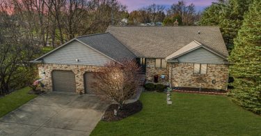 A single-story brick ranch house with a gray shingled roof, two garage doors, a concrete driveway, and a green lawn.