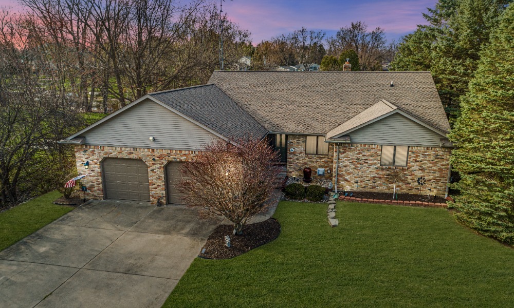 A single-story brick ranch house with a gray shingled roof, two garage doors, a concrete driveway, and a green lawn.