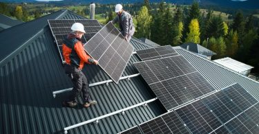 Workers on the roof of a house, placing solar panels on an already installed pipe. There are trees in the distance.