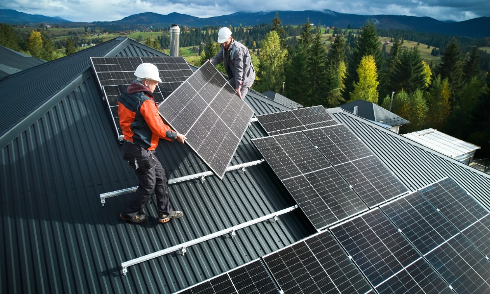 Workers on the roof of a house, placing solar panels on an already installed pipe. There are trees in the distance.