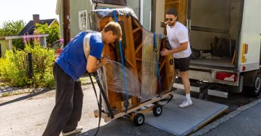 Two movers loading a large, plastic-wrapped piano secured with straps onto a moving truck using a wheeled dolly and loading ramp.