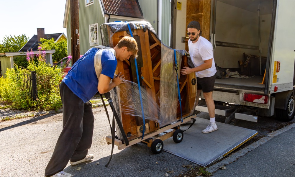 Two movers loading a large, plastic-wrapped piano secured with straps onto a moving truck using a wheeled dolly and loading ramp.