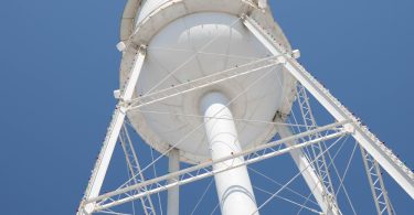 A tall white water tower seen from beneath, with metal support beams and small colored lights against a clear blue sky.