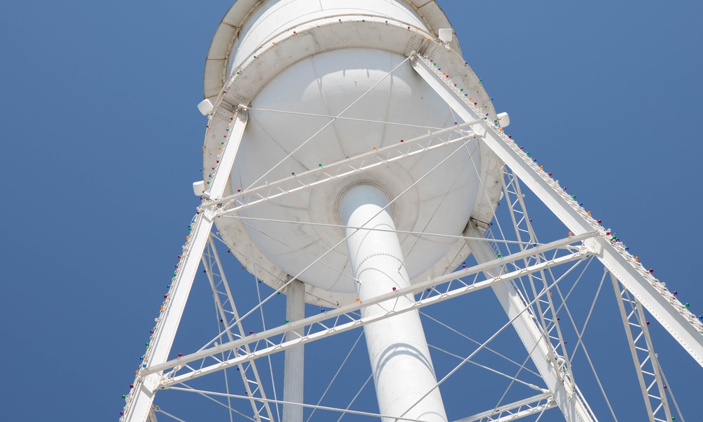 A tall white water tower seen from beneath, with metal support beams and small colored lights against a clear blue sky.