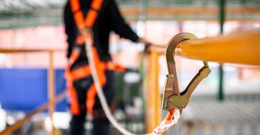 A person wearing an orange safety harness connected by a white rope to a metal hook attached to a yellow railing.